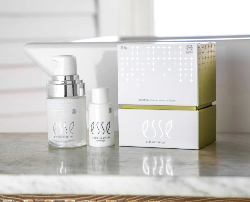 Two bottles of Esse Probiotic Serum and sachets sit on a marble countertop beside white and green box packaging, with a blurred background showing a window and white blinds.